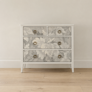 A three-drawer chest of drawers with a white frame and floral-patterned drawer fronts in gray and white, featuring round brass pulls, set against a neutral beige wall and light wood floor.