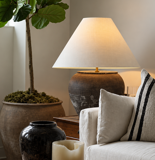 A cozy living room scene with a cream upholstered sofa, textured throw pillows, a lit table lamp on a side table, a potted plant, and a coffee table holding books, vases, and a candle.
