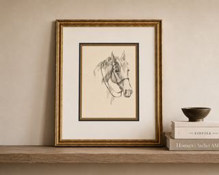 A framed pencil sketch of a horse's head, displayed on a wooden shelf next to stacked books and a small bowl against a beige wall.